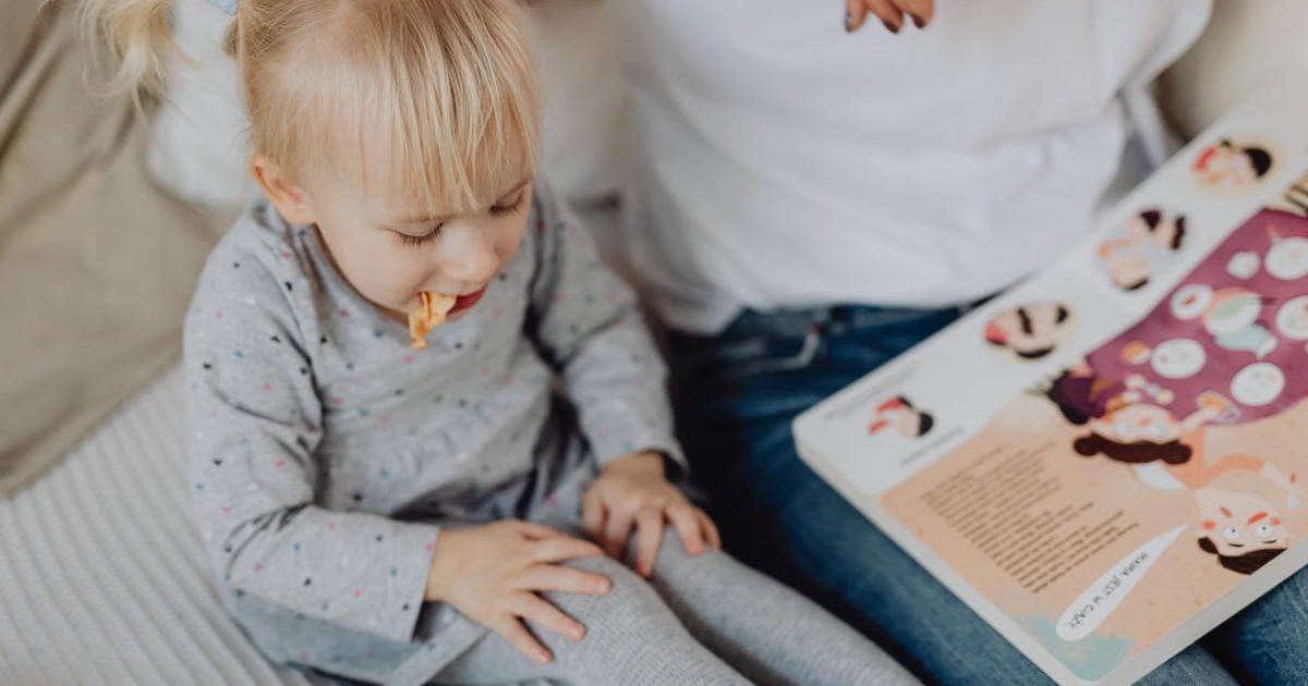 Cute toddler with ponytails enjoying a snack next to a book and parent. Cozy indoor scene.