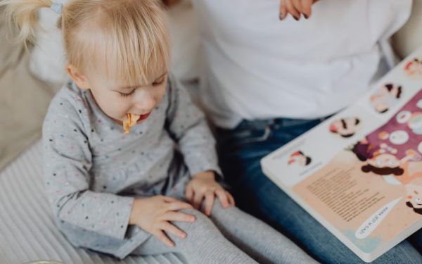 Cute toddler with ponytails enjoying a snack next to a book and parent. Cozy indoor scene.